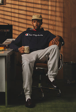 Man in black Champion hoodie and gray sweatpants holding a coffee mug, posed sitting on a desk chair