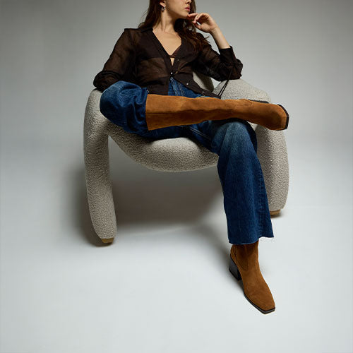 Woman in a dark brown top and jeans with suede brown boots sitting on a white chair