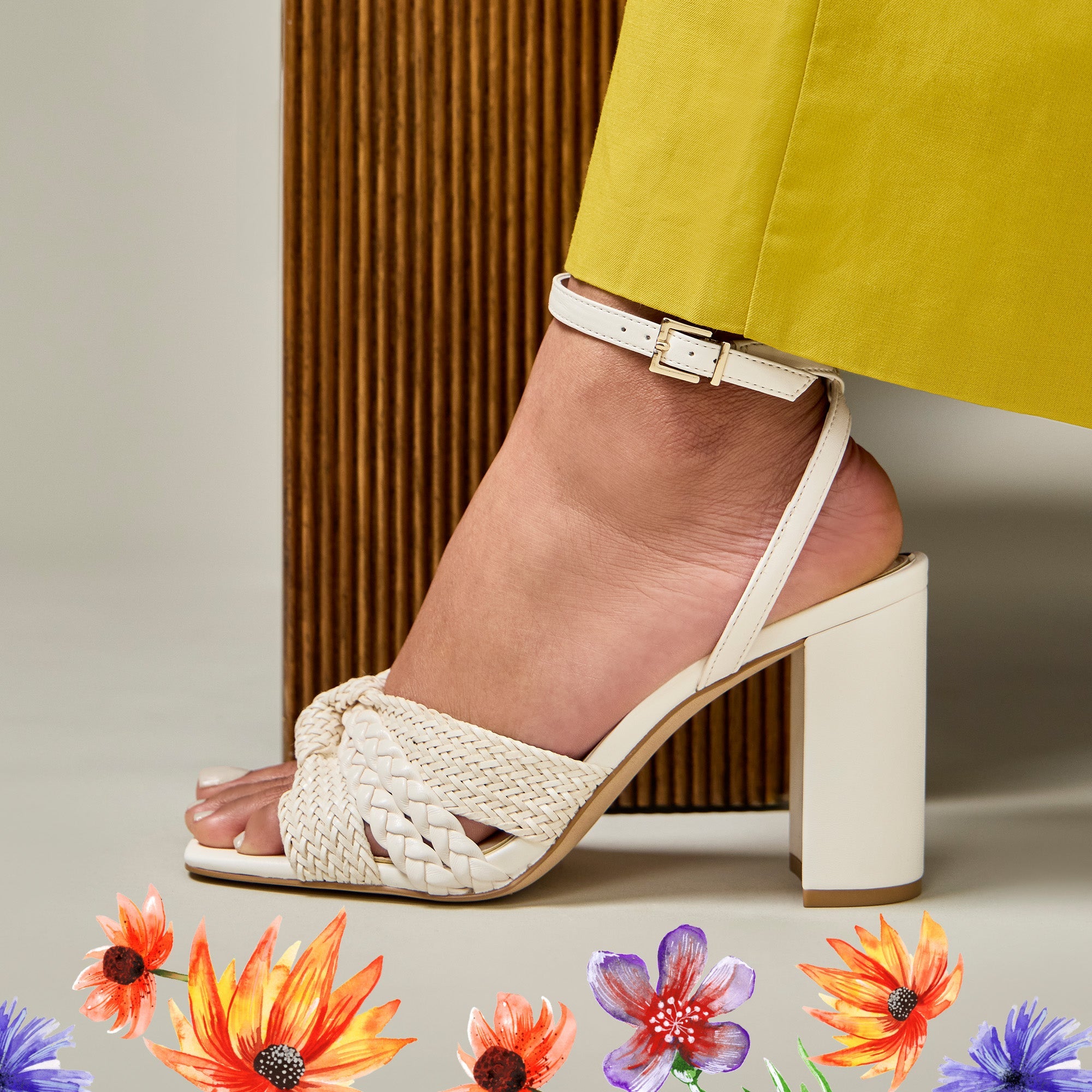 Beige sandal with a block heel worn by a person in yellow pants, with decorative flowers at the bottom.