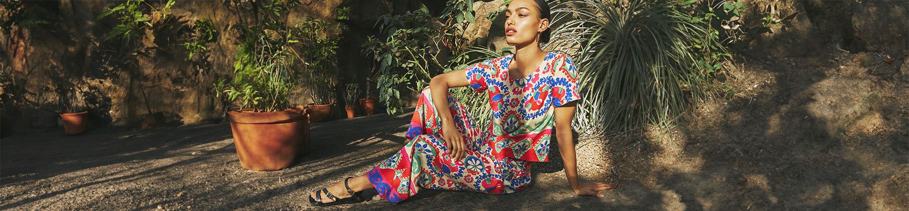 Woman in colorful outfit and black sandals sitting outside surrounded by plants