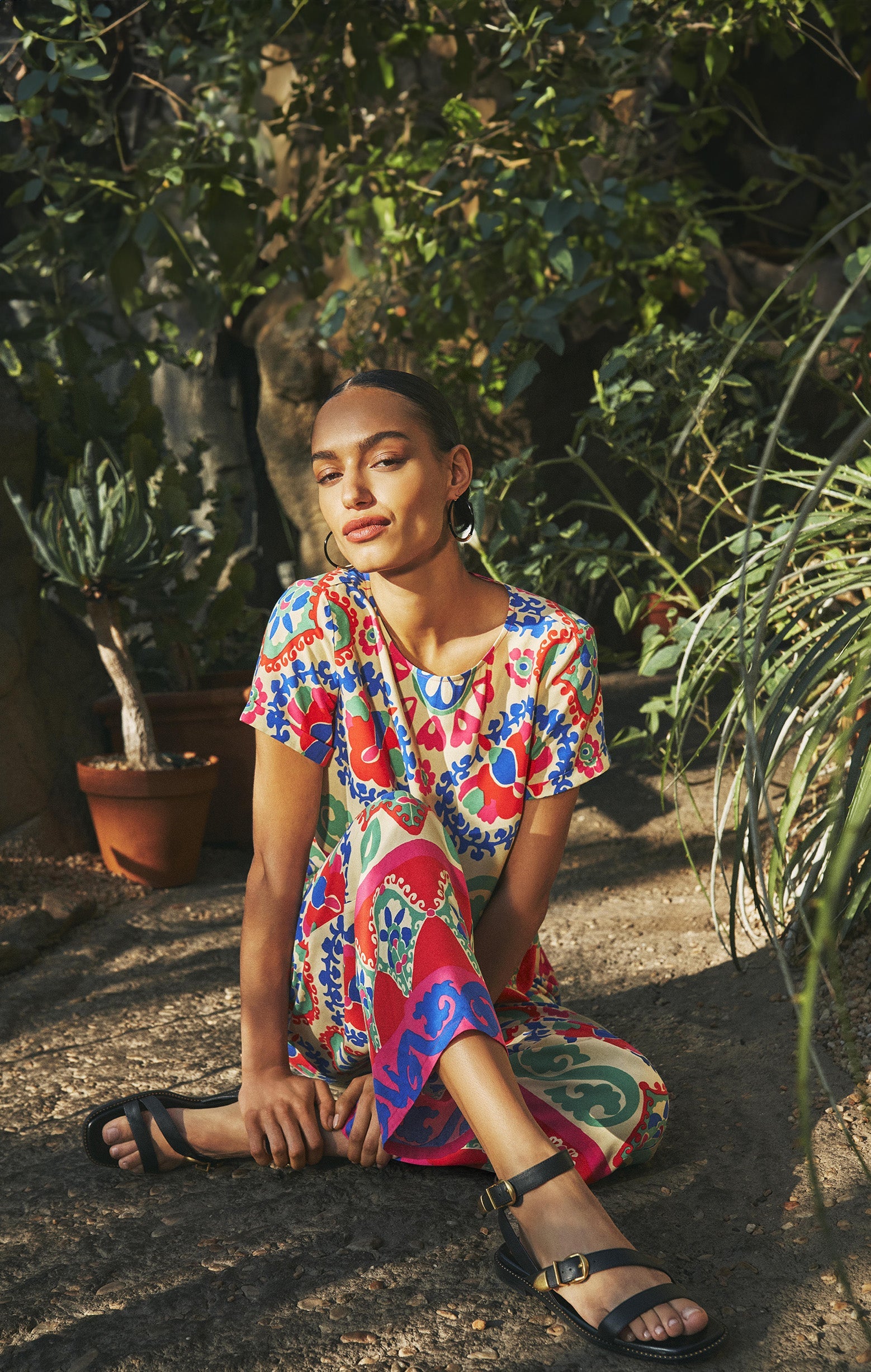 Woman in colorful outfit and black sandals sitting outside surrounded by plants