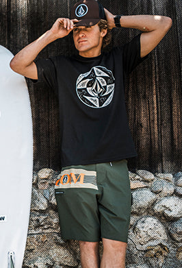 Main adjusting baseball cap, wearing a black shirt and green shorts, while leaning against rocks outside.