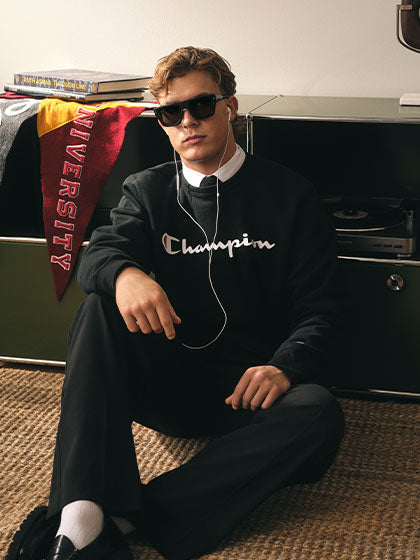 Man in black "Champion" logo sweater with black pants, leaning against file drawers with collegiate materials