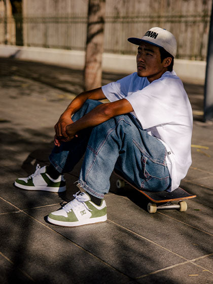 Man in white shirt, jeans, and green and white sneakers sitting on a skateboard outside