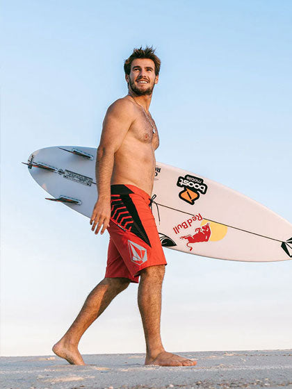 Manin orange swim trunks carrying a surfboard and walking on sand
