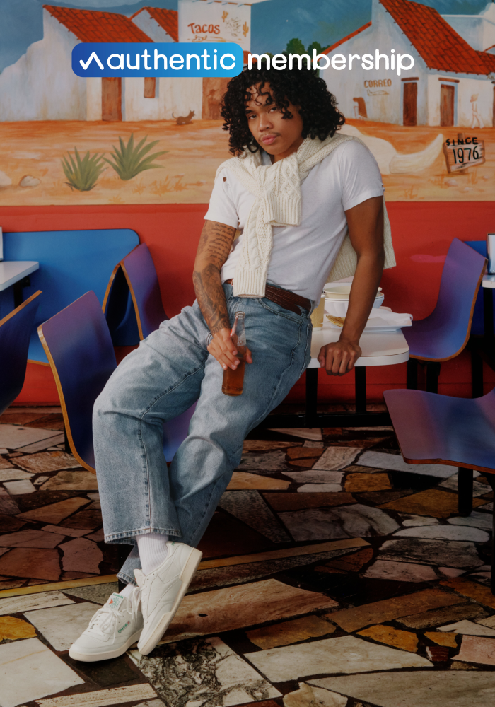 Person in white t-shirt, jeans, and white sneakers leaning against table inside colorful restaurant with an "Authentic Membership" logo at the top