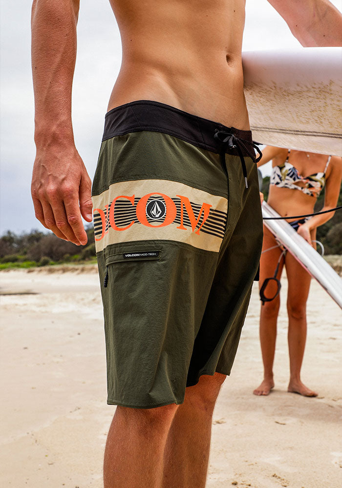 Person in green shorts with Volcom logo, holding a surfboard on the beach