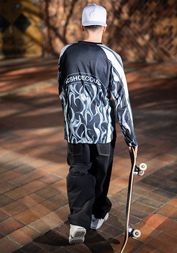 Person in white hat, black shirt with chrome details, and black pants holding a skateboard outside