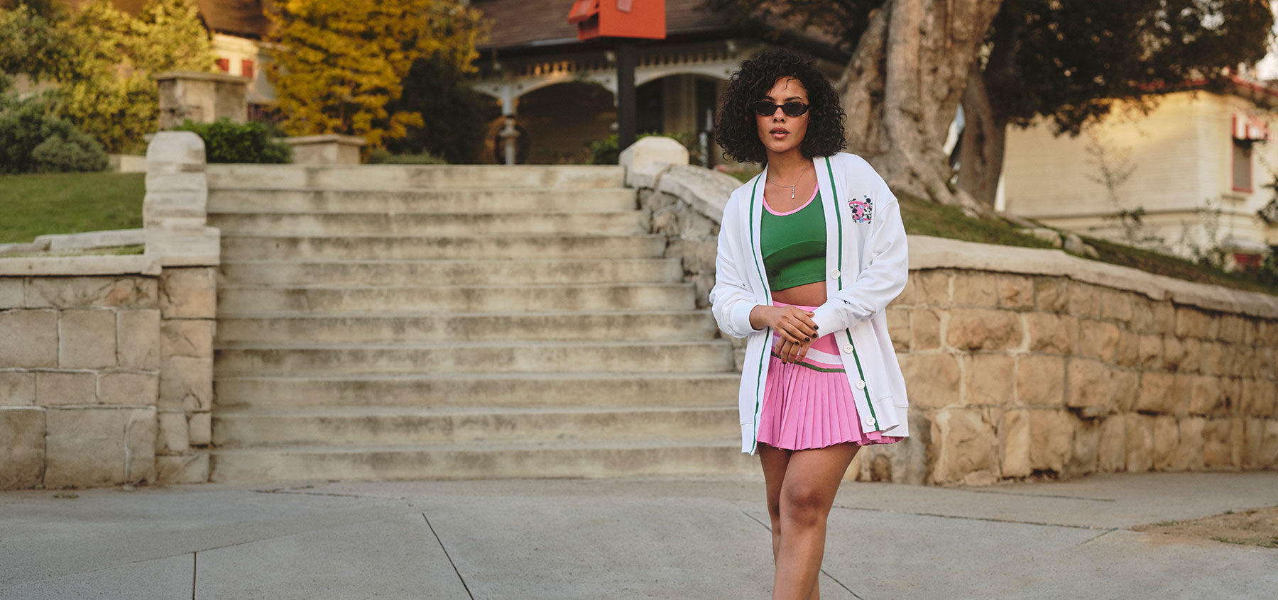 Woman in a white cardigan, green top, and pink skirt standing in front of a staircase outside