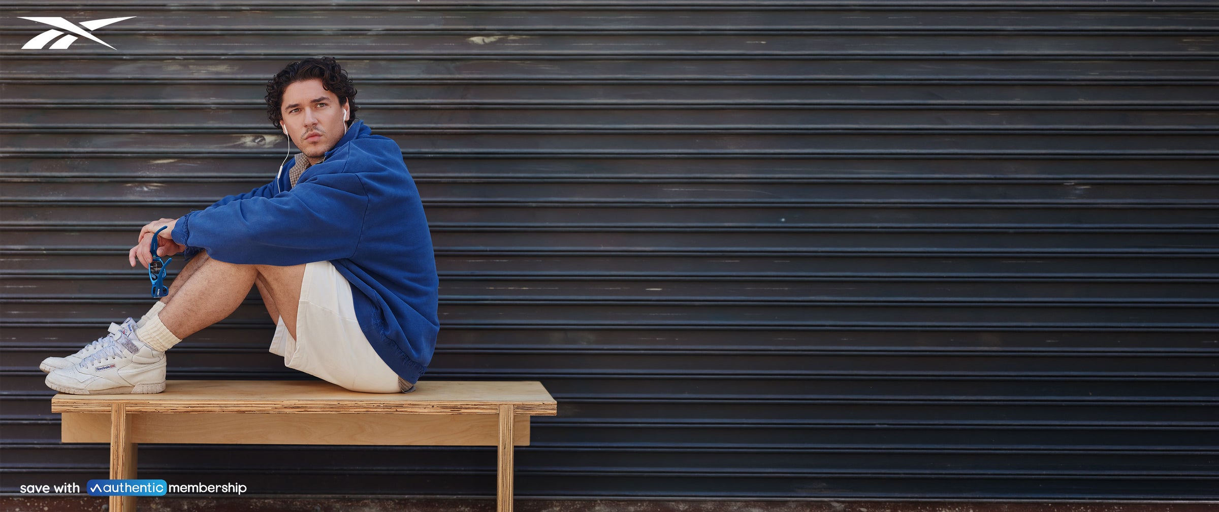 Person in blue sweater, white shorts, and white sneakers sitting on a bench outside. Reebok logo in the upper left and the bottom left has text that reads: "Save with Authentic Membership."