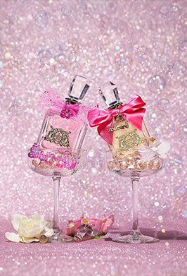 Two perfume bottles with pink bows placed on champagne glasses in front of a pink background filled with bubbles