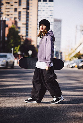 Child in purple hoodie, black pants, and black and white sneakers holding a skateboard crossing a road