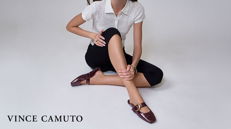Close-up of person in dark red flats sitting on a plain gray studio background with the logo "Vince Camuto" in the bottom left