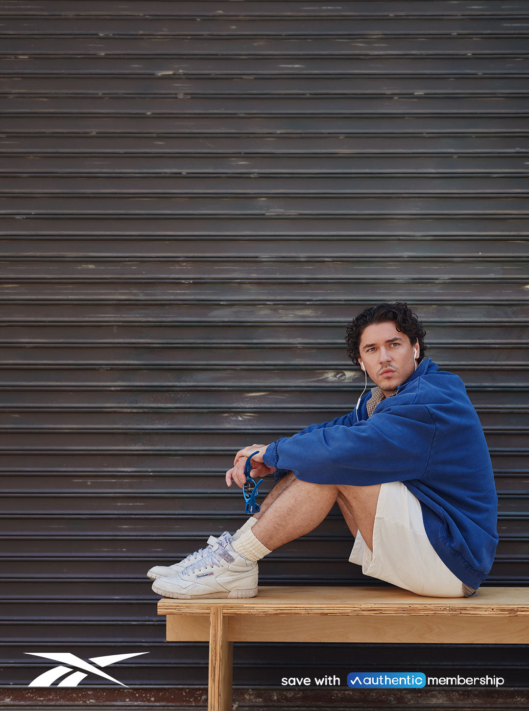 Person in blue sweater, white shorts, and white sneakers sitting on a bench outside. Reebok logo in the upper left and the bottom right has text that reads: "Save with Authentic Membership."