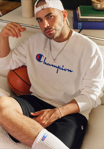 Man posing on a white chair with a basketball in a white sweater with Champion logo on the front.
