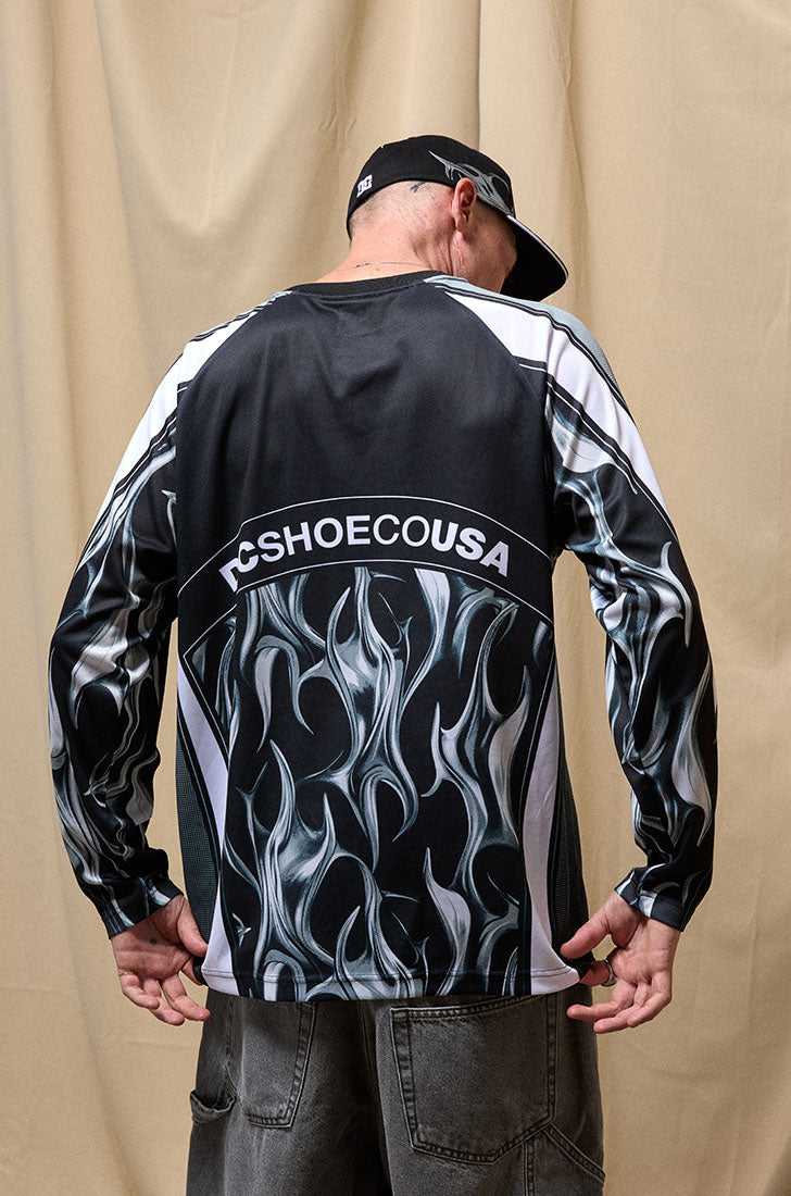 Man in a black cap and black long sleeve shirt with chrome motifs standing against a beige curtain